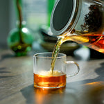 Tea being poured from a glass teapot into a clear glass mug on a wooden surface.