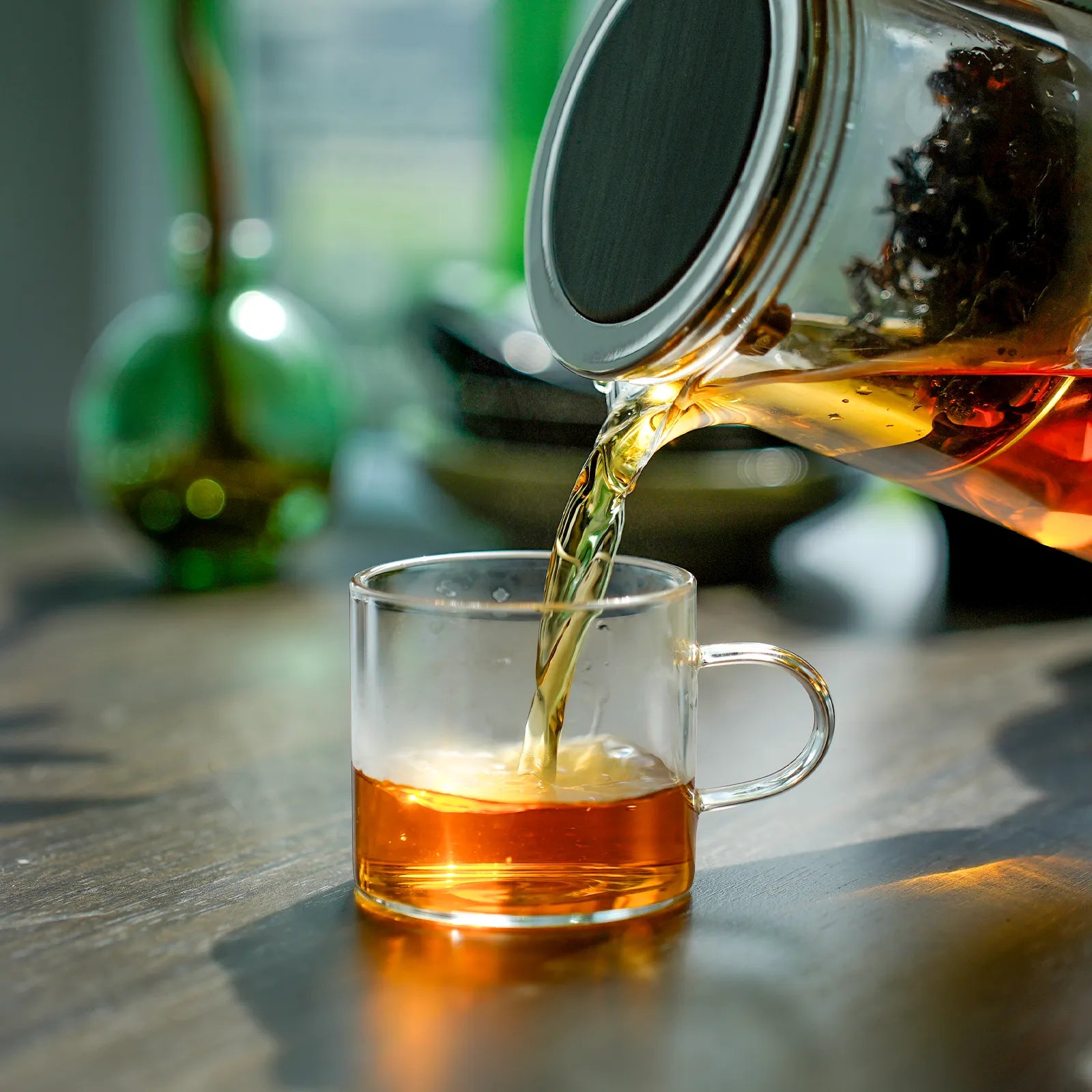 Tea being poured from a glass teapot into a clear glass mug on a wooden surface.