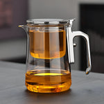 Clear glass teapot with a metal handle and lid filled with orange liquid on a dark surface.