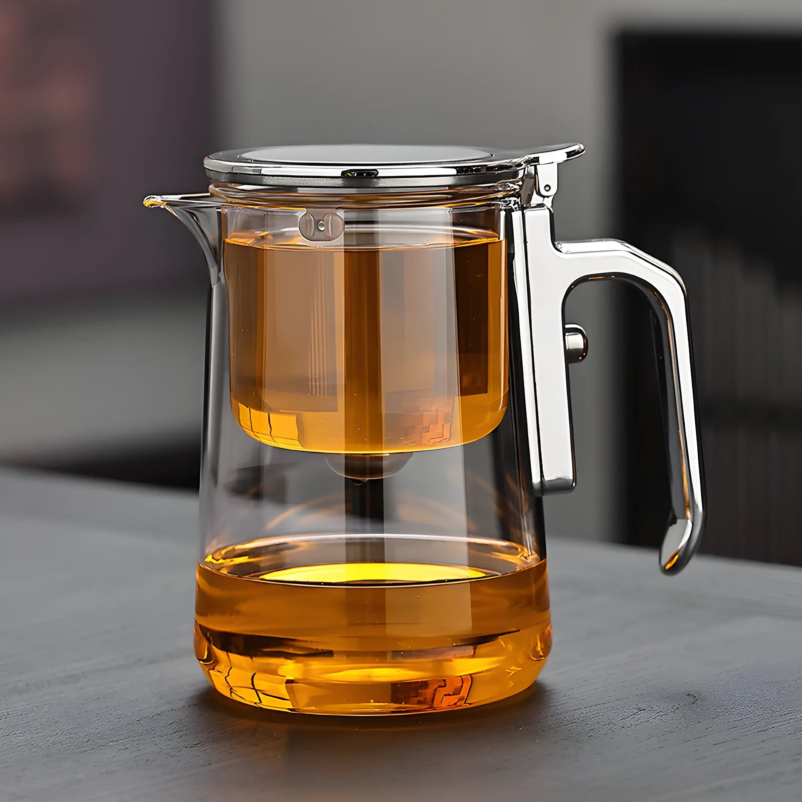 Clear glass teapot with a metal handle and lid filled with orange liquid on a dark surface.