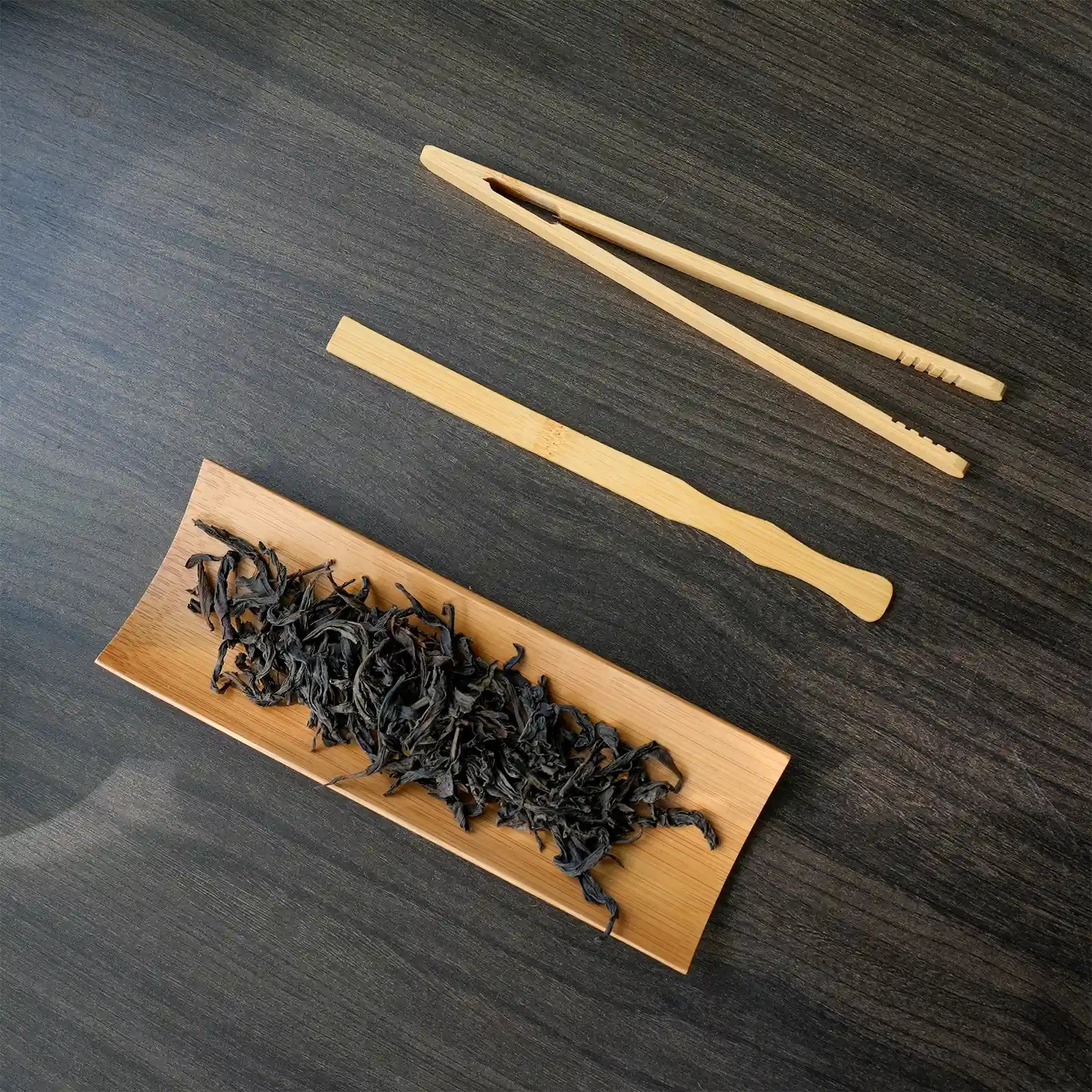 Bamboo tea tongs and a wooden tray with tea leaves on a dark wooden surface